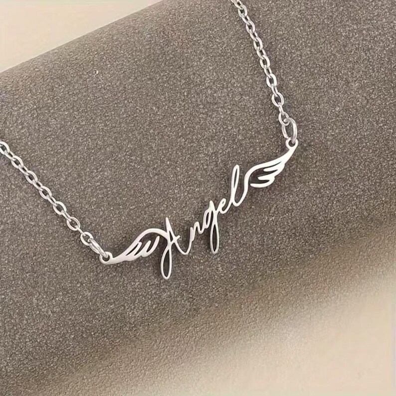 May include: A silver-colored necklace with the word "Angel" in cursive script, flanked by wing designs. The chain is delicate and links together. The necklace is displayed on a gray surface.
