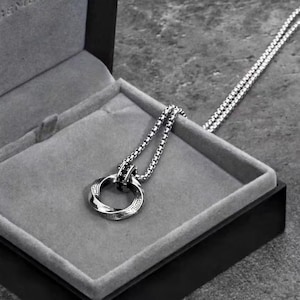 May include: A silver-toned necklace with a circular pendant featuring an engraved design and black accents. The necklace is displayed in a gray velvet-lined box with a black exterior, set against a gray background.