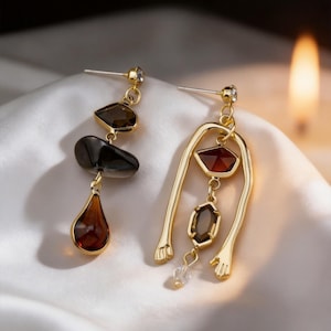 May include: A pair of gold-toned earrings. One earring features a series of dark brown and black stones, culminating in a teardrop shape. The other earring has a unique design with a red gem and a dangling clear crystal.