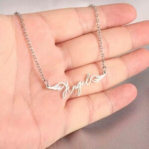 May include: A silver-colored necklace with the word "Angel" in a cursive font. The word is flanked by wing designs. The necklace is held in a person's hand.