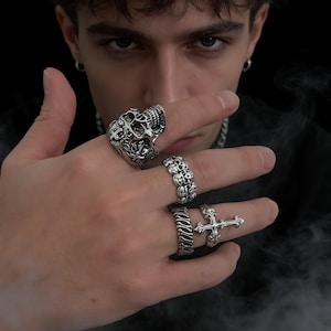 Op de afbeelding: Een hand die een verzameling zilveren ringen toont. De ringen hebben verschillende ontwerpen, waaronder een schedel, schedels, een getextureerde band en een kruis. De ringen worden op meerdere vingers gedragen, wat de ingewikkelde details en stijlen laat zien.