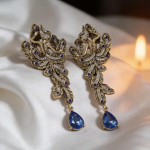 Puede incluir: Un par de pendientes dorados de estilo antiguo. Cada pendiente presenta una figura estilizada, adornada con piedras preciosas claras y azules, y un colgante azul en forma de lágrima. Se presentan sobre una tela blanca y sedosa.