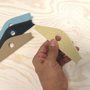 May include: A hand holds a yellow pottery rib tool with a circular hole and a notched edge. Three other ribs in blue, black, and tan are arranged on a light wood surface. These tools are used for shaping clay.