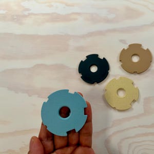 Puede incluir: Cuatro objetos circulares de colores con un agujero central y bordes dentados. Uno se sostiene en una mano. Los colores incluyen azul, negro, beige y amarillo. Sobre una superficie de vetas de madera clara.
