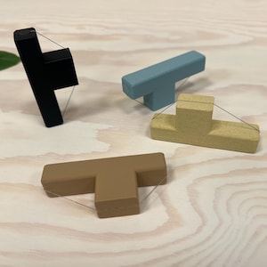 Op de afbeelding: Vier T-vormige houten objecten in zwart, blauw, bruin en beige. Elk heeft een dun draadje bevestigd. De objecten zijn gerangschikt op een licht houtnerfoppervlak. De objecten zijn waarschijnlijk decoratief of functioneel.
