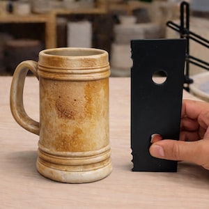 May include: A ceramic beer stein with a handle, featuring a light brown and tan mottled glaze. A black tool with a hole and jagged edge is held nearby. The background shows a pottery studio.