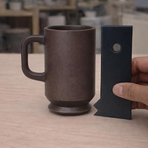 Könnte beinhalten: Eine braune Keramiktasse mit Henkel und ein dunkelblaues Werkzeug mit kreisförmigem Detail. Die Tasse ist zylindrisch mit leicht ausgestelltem Boden. Das Werkzeug ist rechteckig mit einer gezahnten Kante.