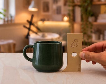 Puede incluir: Una taza de cerámica verde oscuro junto a una herramienta de alfarería de madera color canela claro. La herramienta presenta un dibujo lineal de un gato, un recorte circular y el texto 120 mm impreso verticalmente indicando su altura.
