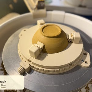 Pode incluir: Um mandril de torno de cerâmica bege autocentrante com três mandíbulas ajustáveis segurando uma tigela de cerâmica amarela invertida em um torno. O dispositivo inclui o texto Sture Studios Trim Chuck Self-centering 3 jaws Fits 25 cm bat-pin wheels.