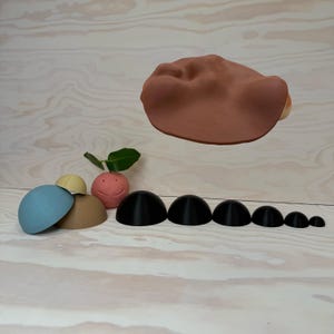 May include: A collection of colourful, semi-spherical objects, including blue, tan, and yellow, next to a smiling pink radish with green leaves. A large, brown, rounded form floats above, with a row of black domes below.