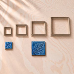 May include: Four square beige plastic cookie cutters in graduated sizes arranged in a row, accompanied by two square blue ceramic tiles featuring embossed fern and leaf patterns, displaying a glossy finish and dark edges.