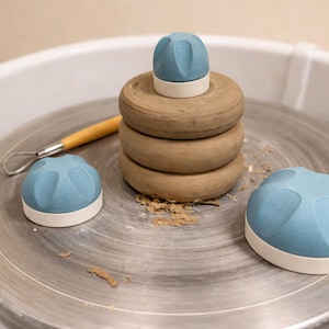 Può includere: Un tornio da vasaio con una pila di anelli di argilla marrone e tre pezzi di ceramica blu e bianca. Uno strumento da vasaio con manico in legno poggia sulla ruota. Gli oggetti sono disposti su una ruota color argento.