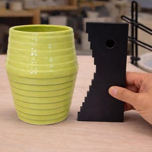 May include: A lime green ceramic vase with horizontal ridges, next to a black pottery rib tool. The vase has a glossy finish. The tool is held by a hand. Pottery tools and equipment are in the background.