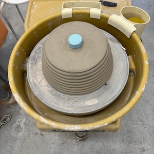 Pode incluir: Uma roda de oleiro com uma tigela de argila castanha clara em andamento. A tigela tem um disco azul claro no topo. A roda é amarela e tem um recipiente de água. Um pequeno banco branco está no fundo.