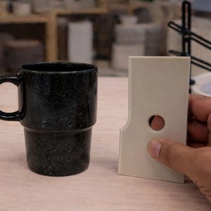 May include: A black speckled ceramic mug with a handle is next to a cream-coloured pottery rib tool. The mug has a cylindrical shape with a slightly flared base. The rib tool has a hole and is held by a hand.