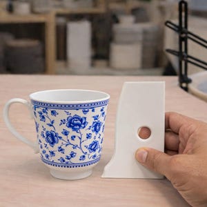 Puede incluir: Una taza de cerámica blanca con un diseño floral azul y una herramienta de costilla de cerámica blanca. La taza tiene un asa y una base. La herramienta de costilla es sostenida por una mano y tiene un agujero circular.