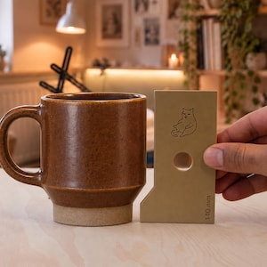 Puede incluir: Una taza de cerámica marrón con acabado moteado y cuerpo cilíndrico, junto a una herramienta de medición beige que presenta un dibujo lineal de un gato, un recorte circular y el texto 140 mm impreso en el borde inferior.
