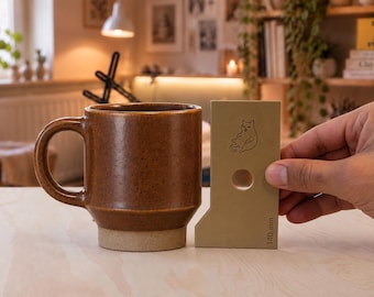 May include: A brown ceramic mug with a speckled finish and a cylindrical body, positioned next to a beige measuring tool that features a line drawing of a cat, a circular cutout, and the text 140 mm printed on the bottom edge.