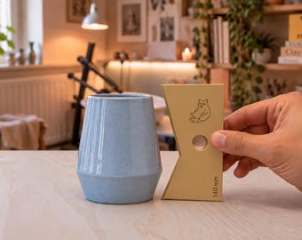 May include: A light blue ceramic cup with a tapered shape stands next to a beige pottery rib tool. The tool features a line drawing of a cat, a circular cutout, and the text 140 mm printed vertically along the right edge.