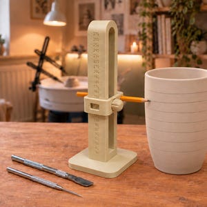 May include: A beige pottery height gauge tool with a vertical scale from 0 to 270 mm, featuring an adjustable horizontal arm holding a yellow pencil. Beside it sits a cylindrical clay vessel with horizontal lines and two metal sculpting tools.
