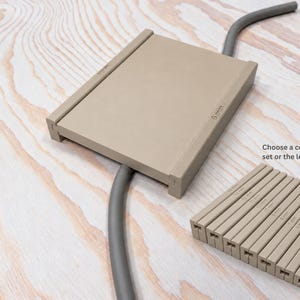 Pode incluir: Uma guia de cabos de plástico bege com uma etiqueta de 6 mm, com um cabo cinzento curvo a passar por ela. Um conjunto de peças guia bege adicionais com várias etiquetas em milímetros está disposto perto, com o texto: Choose a complete set or the length you need.