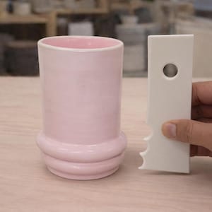 Könnte beinhalten: Ein rosa Keramikbecher mit abgerundetem Boden und ein weißes Keramikwerkzeug mit Loch und gezacktem Rand. Der Becher hat eine glatte, glänzende Oberfläche. Die Gegenstände befinden sich auf einer hellen Holzoberfläche.