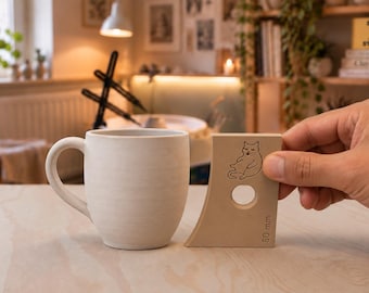 May include: A light-colored ceramic mug with a curved handle positioned next to a tan-colored pottery rib tool. The tool features a circular cutout, a line drawing of a cat, and the text 80 mm printed on the lower right corner.