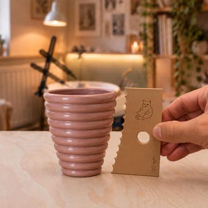 Peut inclure: Une tasse en céramique rose poudré au design côtelé et conique, placée à côté d'un outil de potier rectangulaire beige. L'outil présente un bord dentelé, une découpe circulaire, un dessin de chat et l'inscription 120 mm sur la surface.
