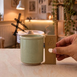 May include: A light green ceramic mug with a smooth finish and a straight-sided cylindrical shape. Next to the mug is a tan measuring tool with a cat illustration and the text 100 mm, indicating the height of the mug is 100 mm.
