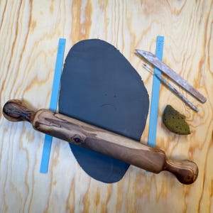 Può includere: Un mattarello di legno con venature scure, una lastra di argilla grigio scuro con una faccia triste e vari strumenti per la ceramica. Gli strumenti includono una spugna, un pennello e uno strumento di legno. Lo sfondo è una superficie di legno chiaro.