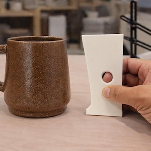 May include: A brown speckled ceramic mug with a handle sits beside a white ceramic tool with a hole. The mug is cylindrical and has a rounded base. The tool is held by a hand.
