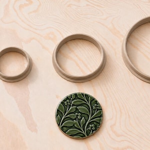 May include: A set of four circular items including two plain beige rings of varying sizes and two circular green ceramic discs featuring a raised botanical leaf and berry pattern. The items are arranged in two rows on a light wood surface.