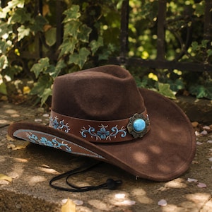 May include: A brown cowboy hat with a decorative band featuring blue floral embroidery. The hat has a turquoise-colored stone accent and a black chin strap. The hat is made of a felt-like material.