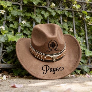 May include: A brown cowboy hat with a decorative band featuring silver studs and a bull skull. The hat has a black emblem and the word "Pages" in script. The hat is resting on a stone surface.