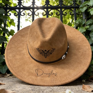 May include: A tan fedora hat with a wide brim. The hat has a black leather band with silver details and a black moth design. The name "Douglas" is written on the brim.