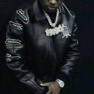 May include: A black leather jacket featuring the word TRAP embroidered in white on the right sleeve. The jacket includes a large silver-toned pendant necklace with the text KRAZY and a shield emblem with the text REX ESTABLISHED 1975.