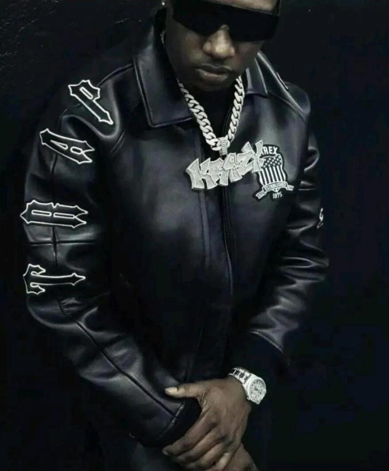 May include: A black leather jacket featuring the word TRAP embroidered in white on the right sleeve. The jacket includes a large silver-toned pendant necklace with the text KRAZY and a shield emblem with the text REX ESTABLISHED 1975.