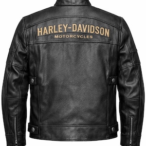 May include: A black leather jacket featuring a mandarin collar, long sleeves with zippered cuffs, and adjustable side tabs with metal snaps. The back displays the text HARLEY-DAVIDSON MOTORCYCLES embroidered in a tan color across the upper panel.