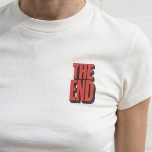 Puede incluir: Camiseta blanca con el texto "THE END" en rojo, delineado en negro, en el pecho. La camiseta tiene mangas cortas y cuello redondo. La imagen es un primer plano de la camiseta.
