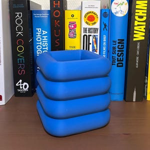 Peut inclure: Une collection de livres aux dos colorés, dont "Watchmen" et "Van Gogh", est disposée derrière un objet bleu en forme de carré. Les livres sont posés sur une surface en bois, créant une présentation visuellement attrayante.