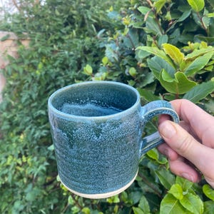 May include: A teal ceramic mug with a textured glaze. The mug has a rounded body, a handle, and a white base. The mug is held in front of green foliage. The mug is suitable for hot beverages.