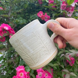 May include: A handmade ceramic mug with an off-white, speckled glaze and a textured leaf pattern embossed on the exterior. The mug features a sturdy, curved handle and a slightly flared rim, showcasing a natural, earthy aesthetic in its design.