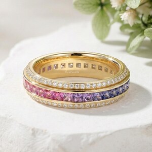 May include: A gold band ring with a row of pink, purple, and blue gemstones. The ring is encrusted with small white gemstones and sits on a white surface. The ring has the text "PuraCaratify".