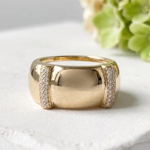 May include: A gold ring with a wide band and two vertical rows of small, clear stones. The ring has a polished, reflective surface and a rounded shape. The ring is set against a white background.