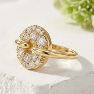 May include: A gold ring with a circular design encrusted with numerous small, sparkling diamonds. The ring features a band that curves around the finger and connects to the diamond-studded circle. The ring is displayed on a white, textured surface.