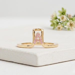 May include: A gold ring featuring a raised, arch-shaped centerpiece that displays the letter A. The letter is embellished with small, round, pink gemstones set into the metal surface, creating a bright and distinct focal point on the band.