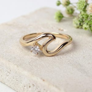 May include: A gold-colored ring with a unique wave design. The ring features a small, round, clear gemstone set in the center of the wave. The ring is displayed on a textured, off-white surface, with soft greenery in the background.