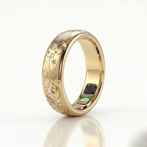 May include: A gold-toned smart ring featuring an engraved pattern of owls, stars, and swirling motifs. The exterior surface is accented with small, clear round stones. The inner band displays two small, rectangular electronic sensor components.