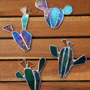 Peut inclure: Quatre ornements en vitrail en forme de cactus, dans des tons de vert, bleu et violet. Chaque cactus est bordé d'argent et posé sur une surface en bois. Les ornements ont un aspect artisanal unique, pour la décoration.