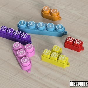 Pu&ograve; includere: Una collezione di tasti di tastiera giocattolo in plastica colorata. I tasti sono in vari colori, tra cui viola, blu, giallo, arancione e rosso. Alcuni tasti hanno lettere, altri numeri. I tasti sono disposti su una superficie chiara con venature del legno.